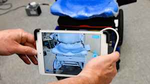 A pair of hands holds a smart tablet to take a digital screening for measurements of foam padding on the seat of a motorized wheelchair