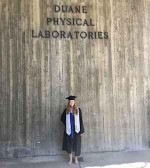A young woman stands in her graduation regalia in front of a building that is named Duane Physical Laboratories on the CU Boulder campus.