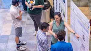 An aerial shot of students standing in front of their posters at the expo. One female points to her poster as two males listen.