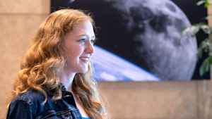 Willow Reed looks to the side as her long red wavy hair falls in front of her jean jacket. On the wall behind her is a large black and white artwork of the moon.