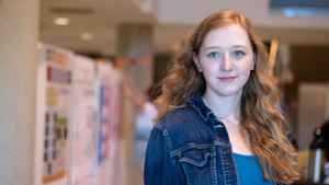 A female astrophysicist looks to the side, her long wavy red hair falling in front of her jean jacket at a student research expo