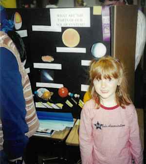 A young redheaded girl in kindergarten stands in front of her science fair poster about the solar system.