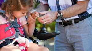 A male occupational therapist takes the hand of a young girl and helps her grab the joystick of a motorized wheelchair. A stuffed pink bunny sits on her lap.