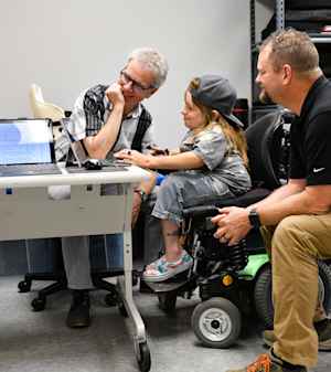 A young woman sits in her motorized wheelchair and works on a laptop as two CU staff members look on and smile. They discuss how to improve her wheelchair.
