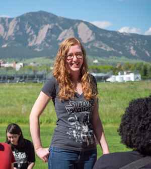 A young woman with long redy wavy hair stands with her hands on her hips and smiles amind the group of college students working on experiments outside. The young woman is wearing a black Star Wars T-shirt and eyeglasses.