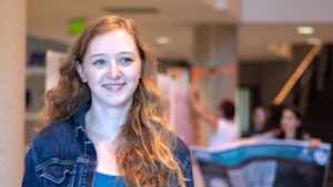 Willow Reed smiles and looks off to the side, her red wavy hair hanging over her jean jacket. She is standing in the middle of an expo where students share posters about their scientific research.