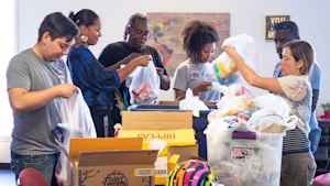 A group of CU students and researchers stand at a table filled with boxes and help community organizers put chips, granola bars and toothbruches into plastic bags to deliver to the neighborhood residents.