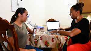 A teenage girl sits at a kitchen table and blows into a spirometer to measure her lung function. A female CU researcher sits across from her looking at the measurements on a computer.