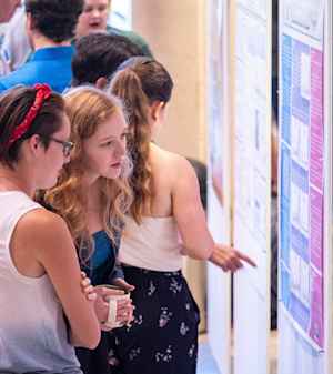 Willow Reed leans in intently to hear a female college student explain her research poster at an expo.
