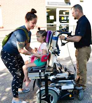 Outside in a parking lot, a mother straps her young daughter into a motorized wheelchair as a clinic staff member makes adjustments.