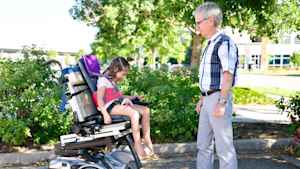 In a parking lot outside the clinic, a young girl gleefully laughs as she moves the motorized wheelchair forward for the first time. A male occupational therapist looks on.