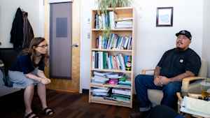 A female mental health clinican leans over in her chair to listen to a veteran sitting across from her during a therapy session. A bookcase filled with books and plants is against the wall.