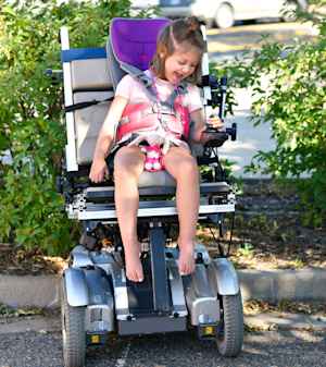 A young girl laughs and looks at her hand around the joystick as she sits in a motorized wheelchair for the first time.