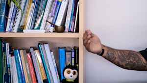 A heavily tattooed arm of a man is extended as the veteran tells how he survived combat in front of a bookcase filled with self-help books, a stuffed animal and other trinkets.