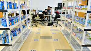 Three male CU students cluster around a laptop at the front of a classroom, where a mock warehouse has been set up with shelves lined with cans and boxes of food. QR codes line the floor.