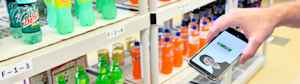 A hand holds a smart phone showing a bottle of soda. The phone's software confirms for the young man that the soda bottle he is looking for is in front of him on the shelf.