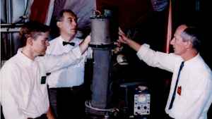 An old historic photo where three male scientists, one with a pocket protector, one with a bowtie and one with a skinny tie, stand around holding an early rocket.