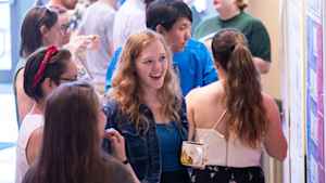 Willow Reed exclaims excitedly with her mouth open in the middle of a crowded student research expo while listening to a female college student explain her work.