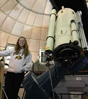 Willow Reed as an undergraduate student at CU stands in front of a large telescope. She is smiling and wearing a white CU Boulder sweatshirt.