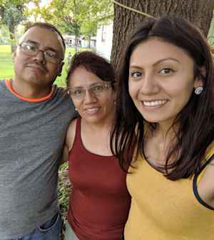 A female CU student stands side by side with her father and mother during a backyard barbecue.