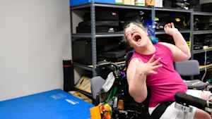 A woman with bright blue hair and wearing glasses throws her head back to laugh as she sits in her wheelchair in the clinic.
