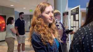 A young woman with long wavy red hair listens intently to a female college student explain her research in a crowded room. They are at an expo where college students share posters about the research they worked on all summer.