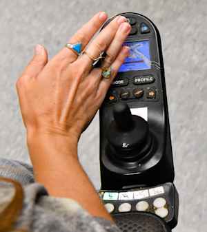 A female's hand wearing rings on three fingers, touches the cracked screen on the joystick of her motorized wheelchair.