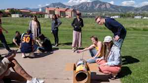 Clusters of college students stand outside on the CU Boulder East campus working together to build different projects. One group is working on a tube that will funnel sunlight. In the forefront a female student with a large leg tattoo checks her phone.