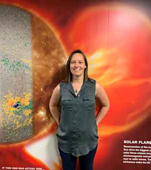 A female scientist stands with her hands on her hips in front of a wall showing red and yellow images about solar flares.