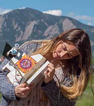 A female college student examines in her hands a solar-powered machine she and her classmates built. On the front of the contraption, which has gears and a mirror, is taped a printout of a cat wearing a sunflower costume. The flatirons are in the background.