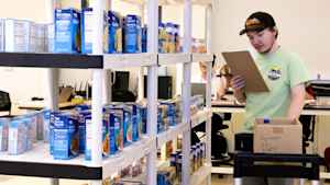 A young man walks holds a clipboard and looks at a shopping list while pushing a cart in a mock warehouse. The shelves are lined with cans and boxes of food.