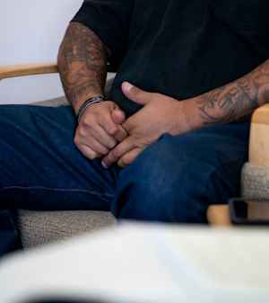 A man's hands clasp tightly together, the arms heavily tattoed, as he sits in a chair