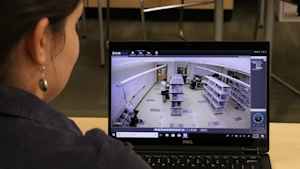 A female CU student watches on a computer screen live video of a client walking through the mock warehouse to monitor his movements.