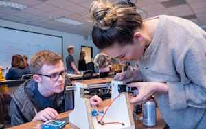 A male college student kneels to get a better look at a solar-powered camera his female classmate is leaning over. She is trying to attach wires and other materials to make it function. They are inside a classroom where other students are also trying to build similar projects.