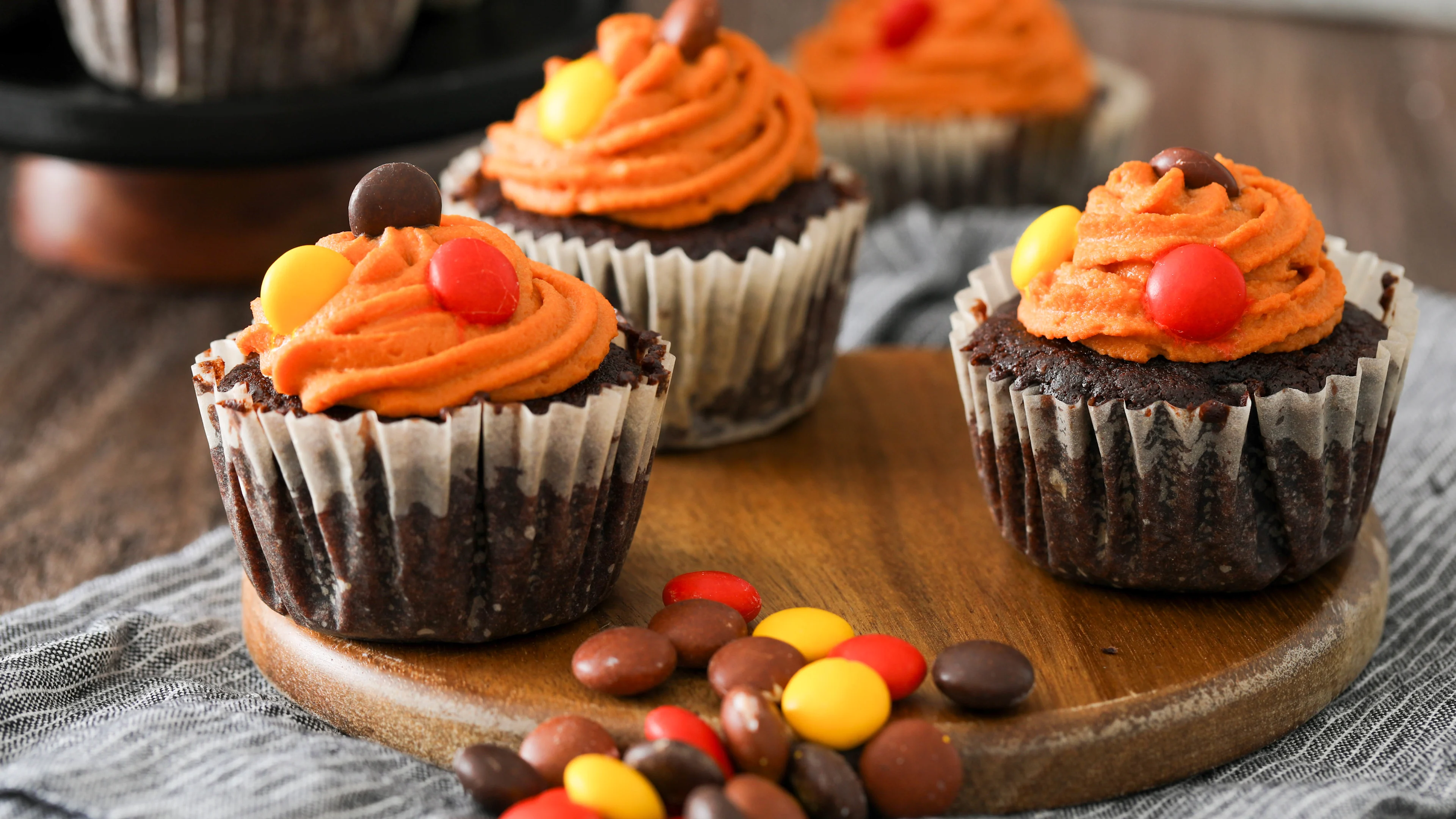 Chocolate cupcakes with orange frosting topped with colorful candy pieces on a wooden board, surrounded by scattered candies.