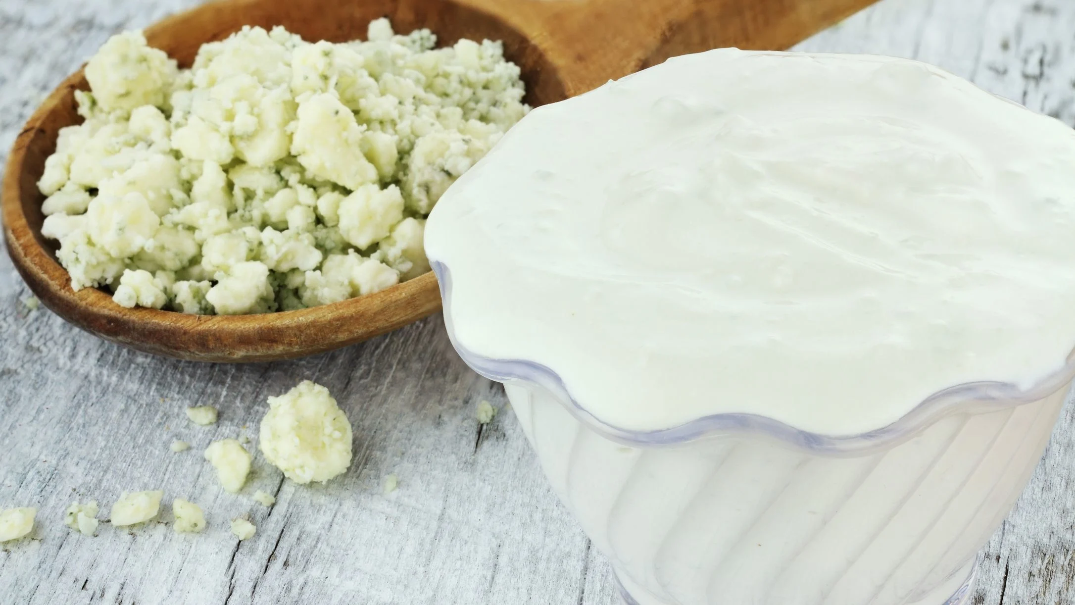 Crumbled blue cheese in wooden spoon next to container of creamy white dressing on weathered wooden surface.