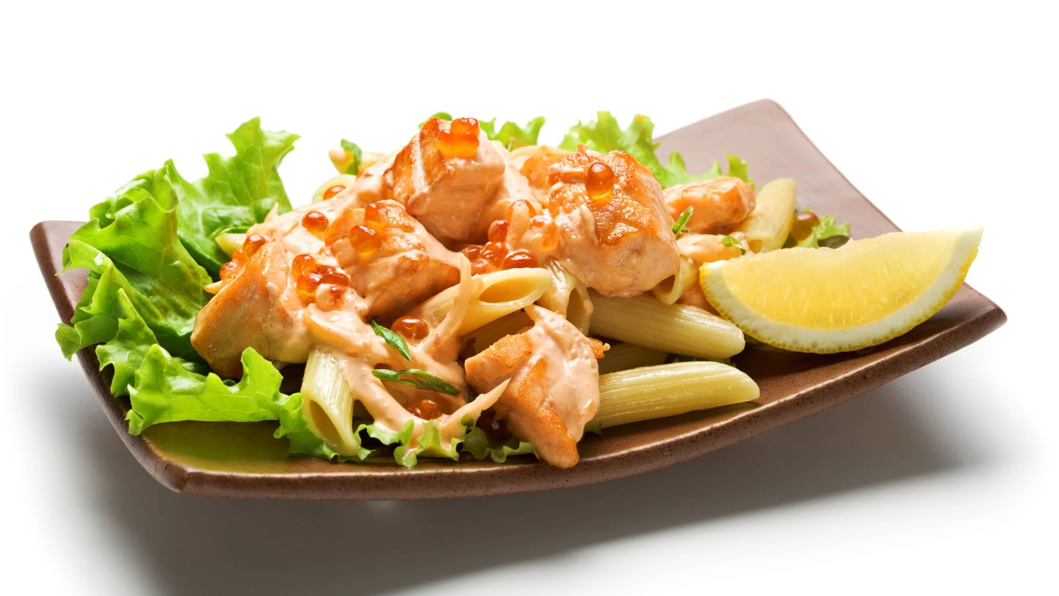 Salmon with creamy sauce over penne pasta, served with fresh lettuce and a lemon wedge on a brown plate.