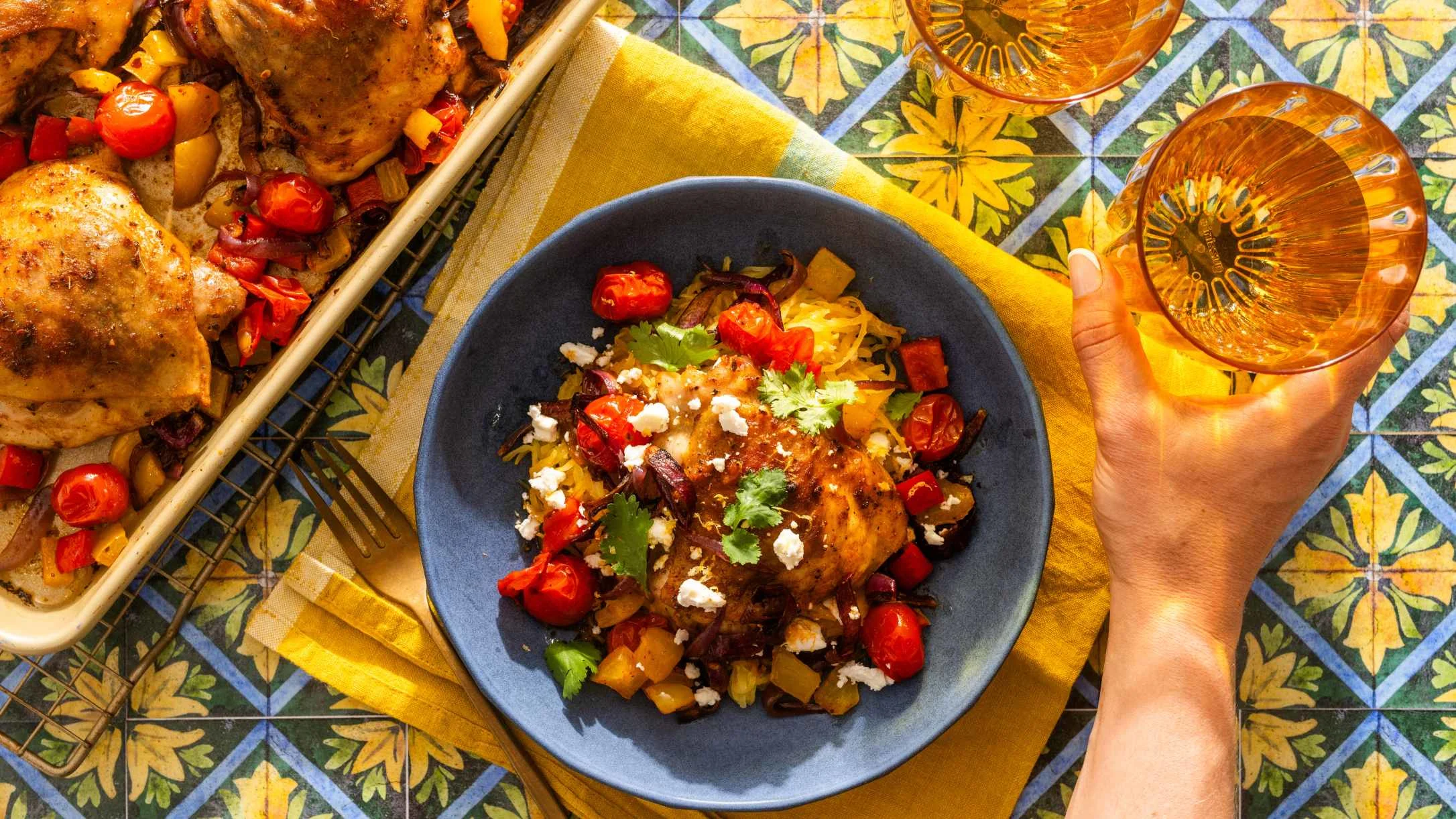 A blue bowl of roasted chicken with tomatoes, peppers, and feta on a colorful tile surface beside a sheet pan meal.