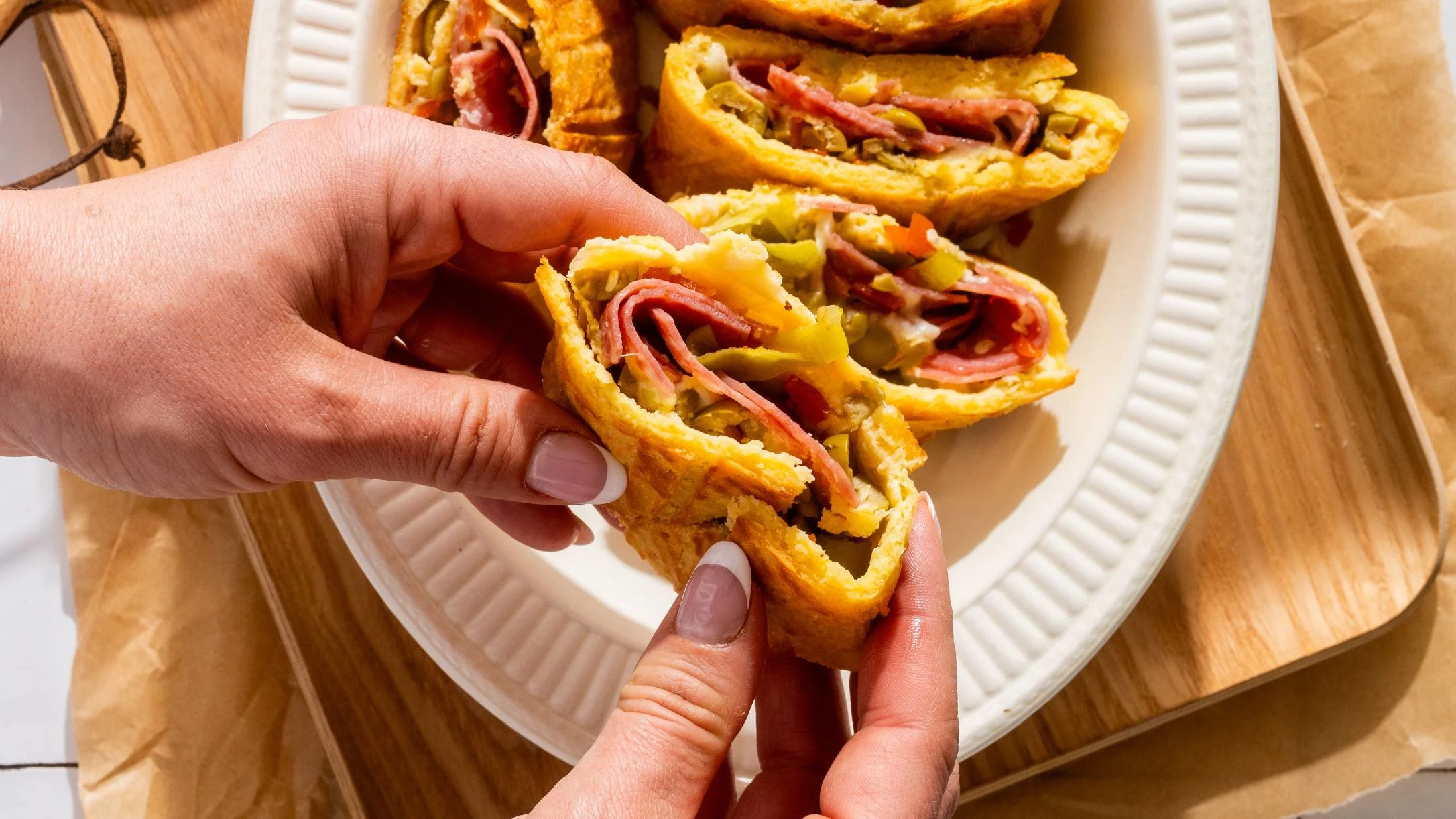 Hand holding a muffuletta-style sandwich wrap filled with ham and pickles on a white plate on wooden surface.