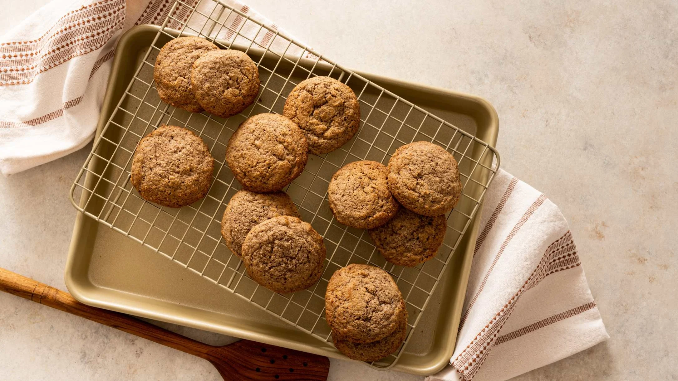 Freshly baked brown cookies cooling on a wire rack over a gold baking sheet, with striped kitchen towels nearby.