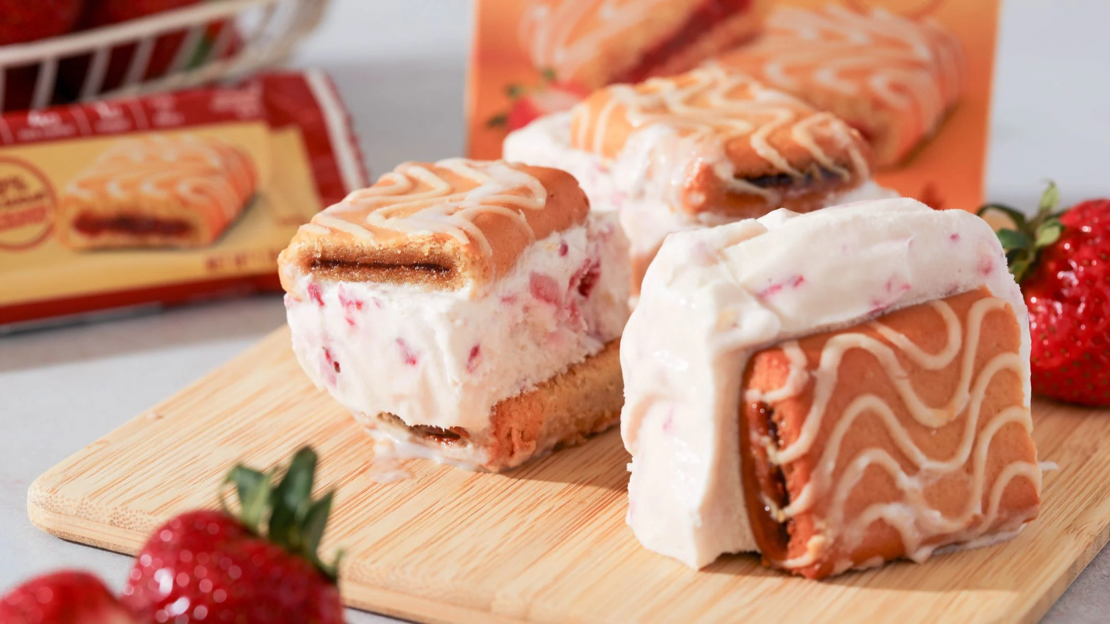 Strawberry ice cream sandwiches with swirled cookie wafers on a wooden board, surrounded by fresh strawberries and product packaging.