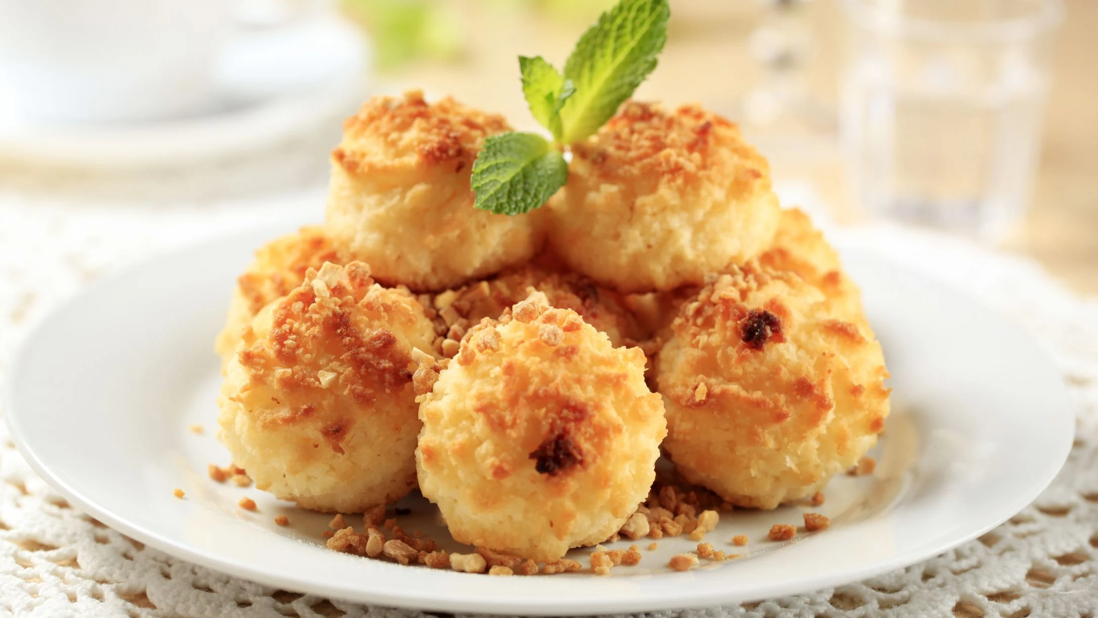 Golden coconut macaroons stacked on a white plate, garnished with fresh mint leaves and scattered crushed nuts.