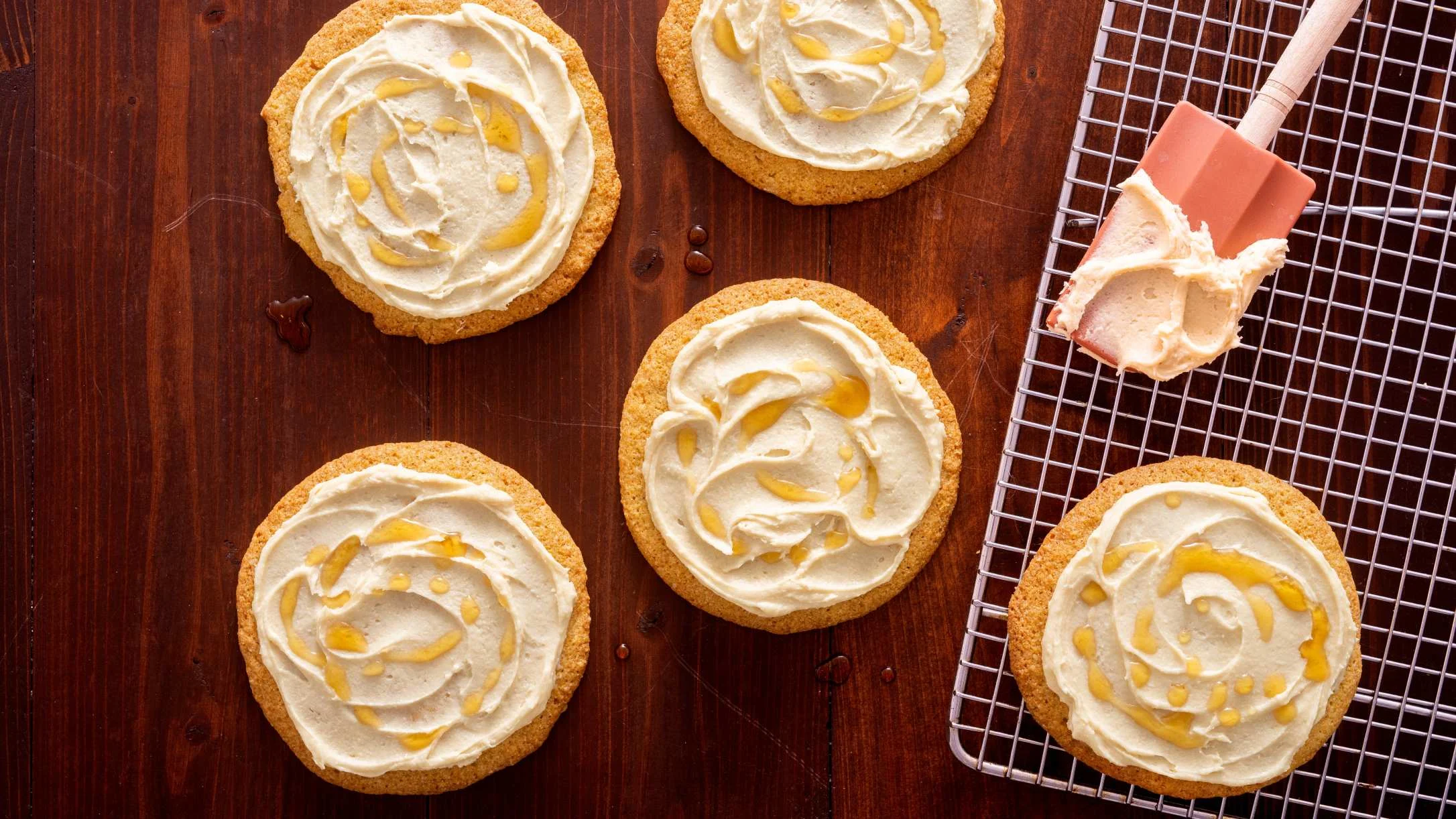 Sugar cookies with cream frosting and drizzle on wooden surface, with spatula and cooling rack visible.