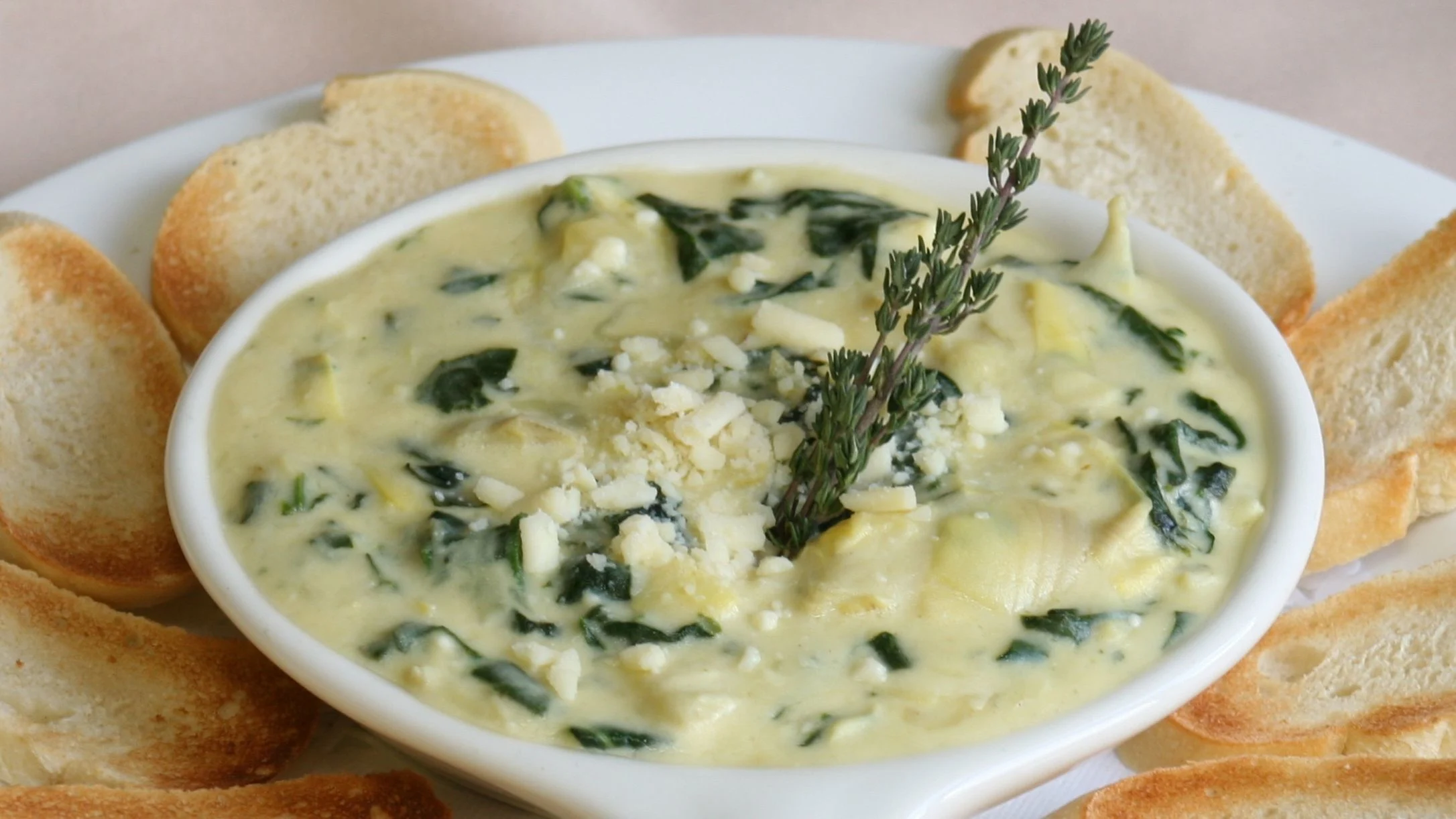 Creamy spinach and artichoke dip in a white bowl, garnished with fresh thyme, surrounded by toasted bread slices.