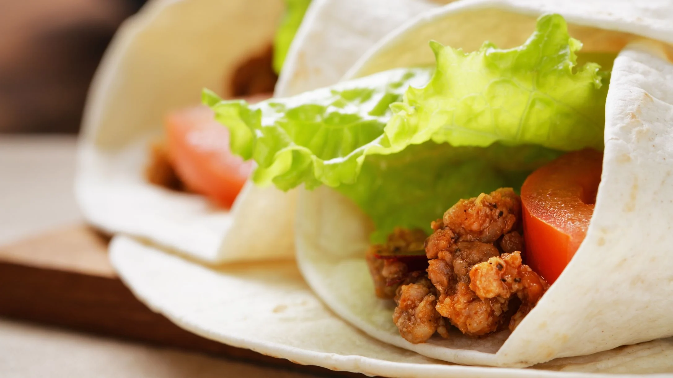 A close-up of a flour tortilla wrap filled with seasoned tofu, fresh tomato slices, and bright green lettuce leaves.