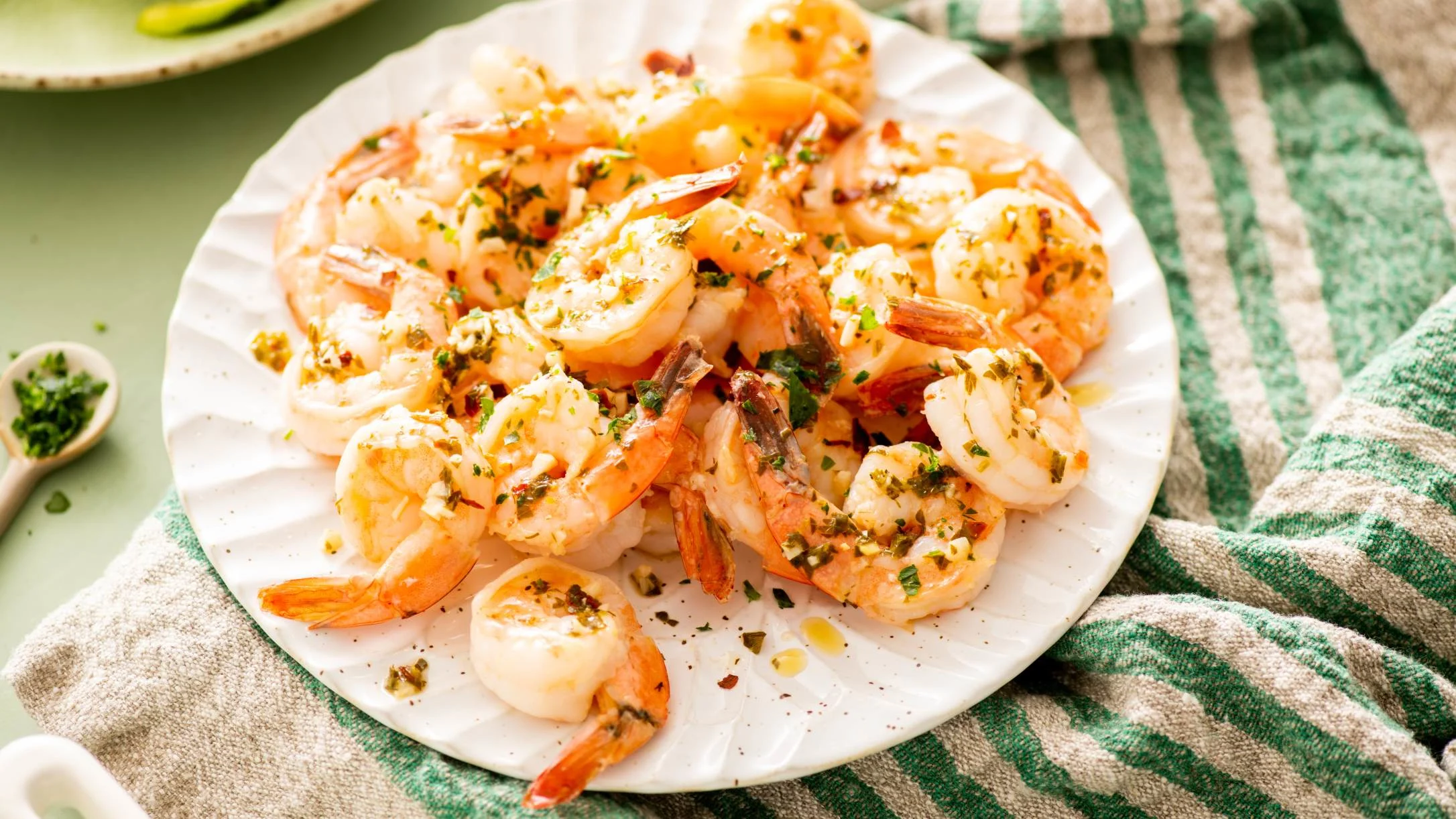 A white plate piled with cooked shrimp topped with garlic, herbs, and butter sauce, set on a green striped cloth.