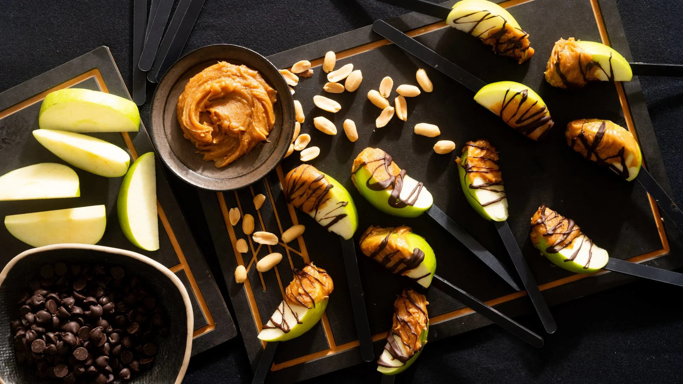 Green apple slices on sticks topped with peanut butter and chocolate drizzle, displayed on black trays with peanuts and chocolate chips.