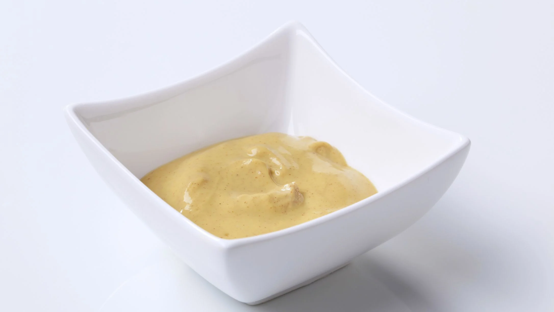 A small white square ceramic bowl filled with creamy yellow mustard sauce on a white background.