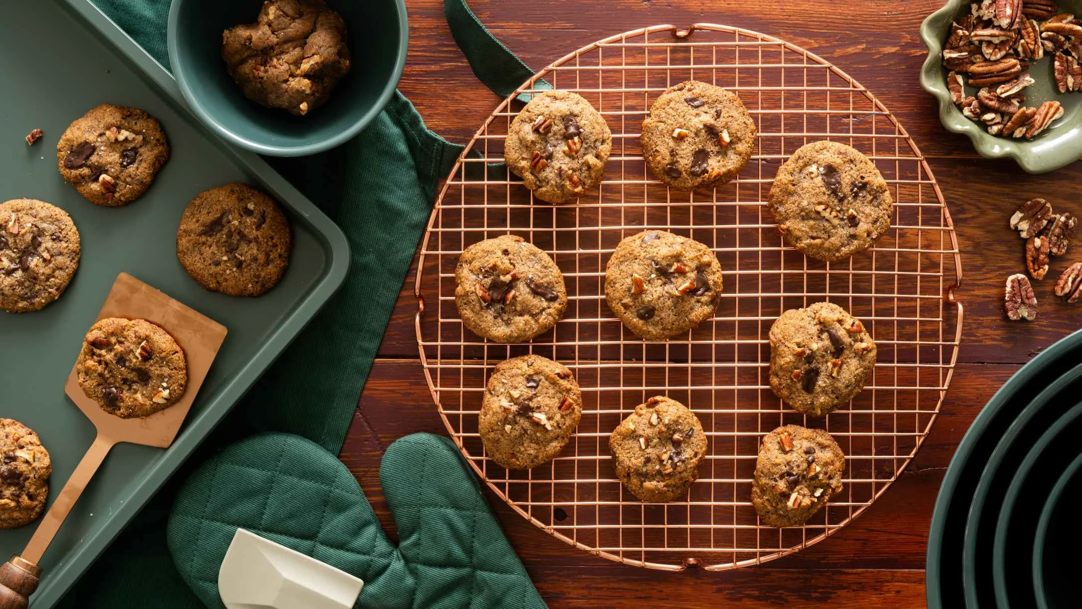 Freshly baked chocolate chip pecan cookies on a copper cooling rack, with more cookies on a green baking tray nearby.