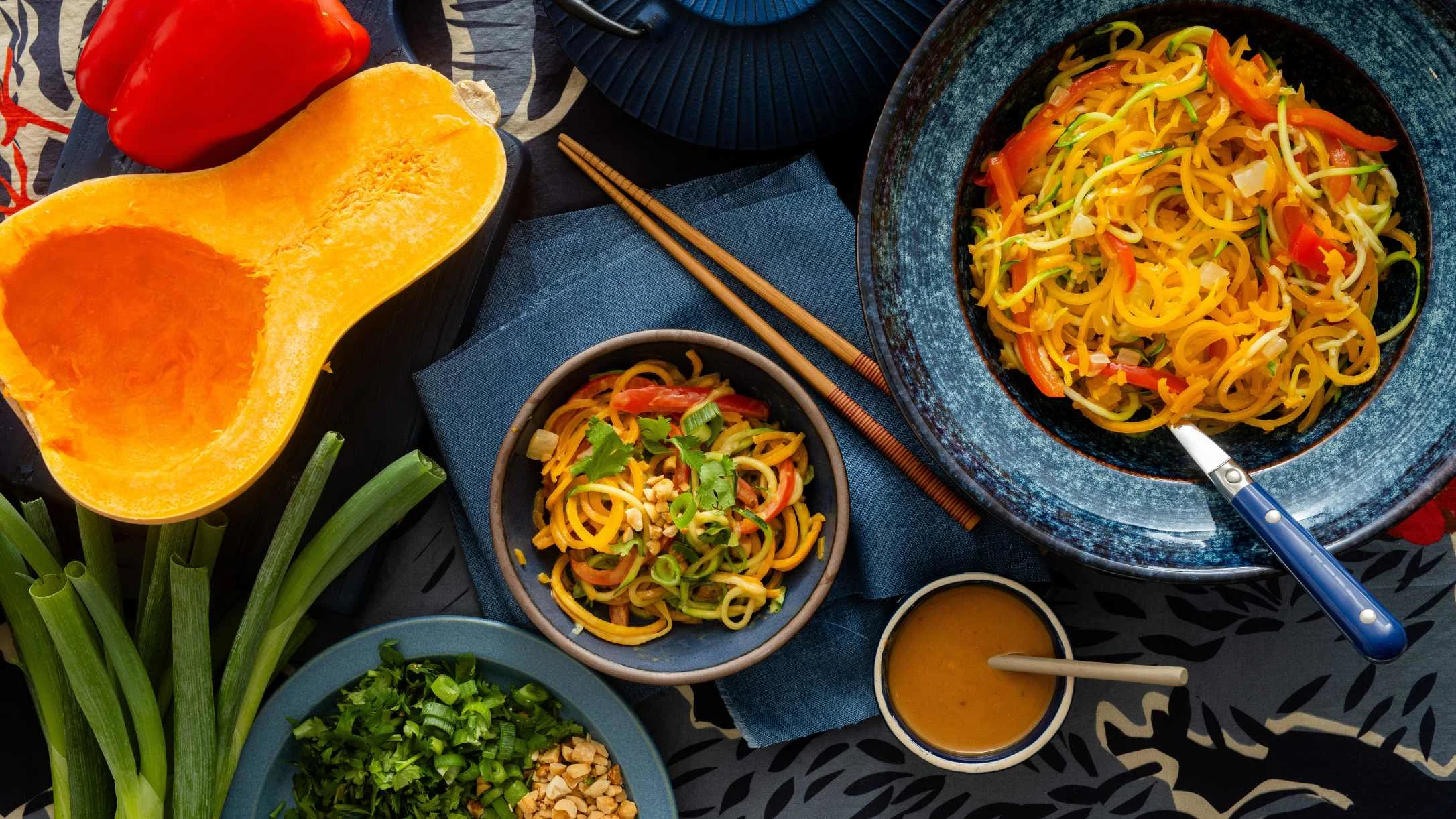 Two blue bowls of spiralized vegetable noodles with red pepper, herbs, and peanut sauce, surrounded by fresh ingredients.